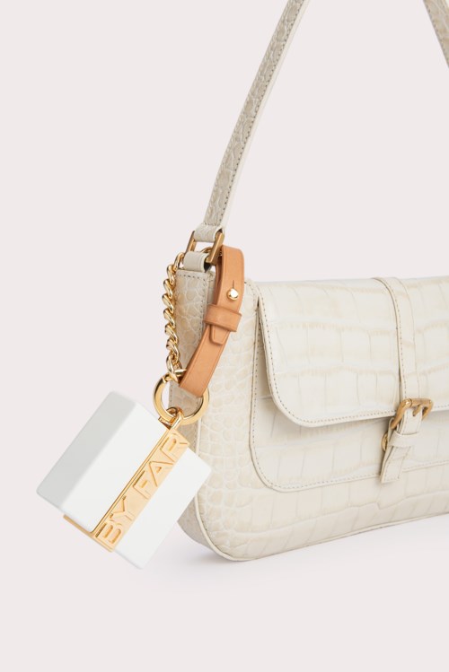 Eine weiße Tasche mit Träger, Goldkette und Schlüsselring vor einfachem Hintergrund.