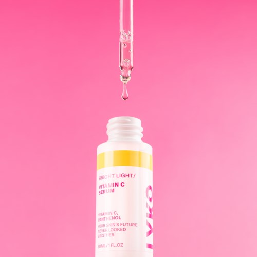 Een pipet brengt serum in een klein wit flesje met een gekleurld label, tegen een roze achtergrond.
