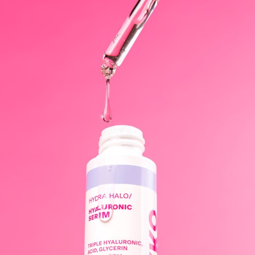 Een roze pipet druppelt serum in een witte fles tegen een roze achtergrond.