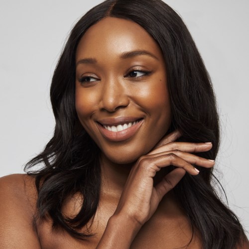 A woman with long black hair smiling gently, touching her face with one hand, wearing a simple top.