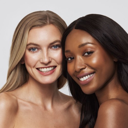 Twee vrouwen met verschillende huidtinten glimlachen dicht bij elkaar in een vriendschappelijke pose.