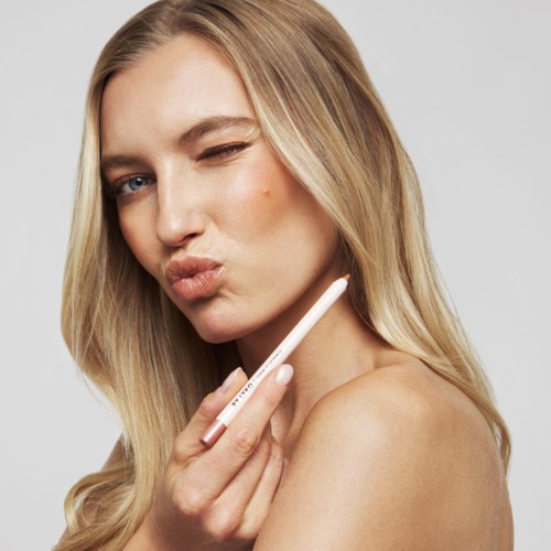 A woman with long blonde hair holds a makeup brush near her face, giving a playful kiss and winking.