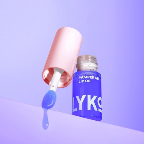 Een blauwe fles met de tekst 'Pamper Me Lip Oil' en een applicator, op paarse achtergrond.
