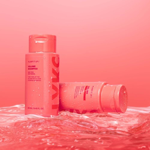 Twee roze shampoo flessen staan op een nat oppervlak met waterreflecties en roze achtergrond.