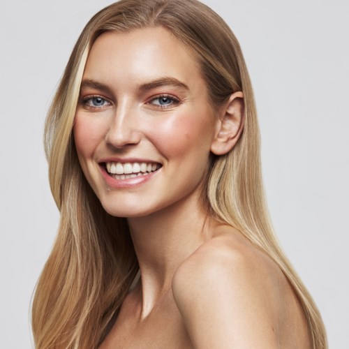 Eine lächelnde junge Frau mit langem blondem Haar und blauen Augen, schaut in die Kamera vor einem schlichten Hintergrund.