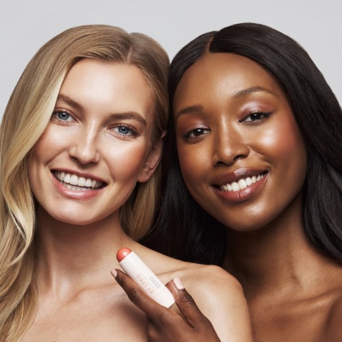 Zwei Frauen lächeln, eine hält Lippenstift, ihre Haut sieht glatt und strahlend aus.