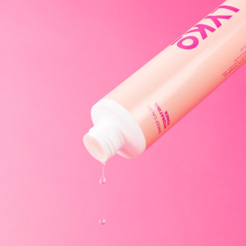 Een roze fles met witte dop die een druppel vloeistof uitgeeft tegen een roze achtergrond.