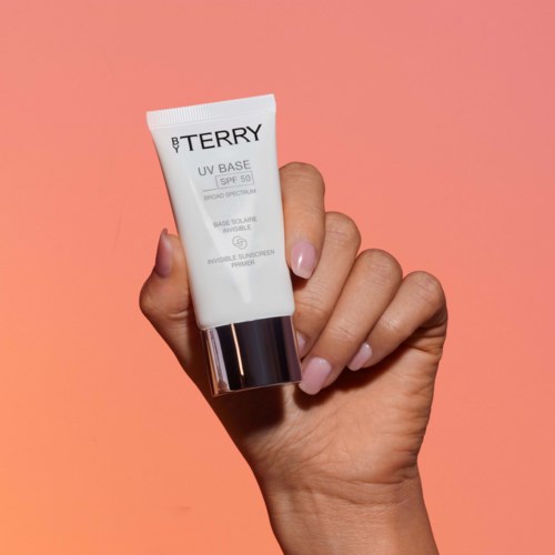 Hånd som holder en tube med UV-krem med rosa bakgrunn, etiketten viser SPF 50 og merkevaren BY TERRY.