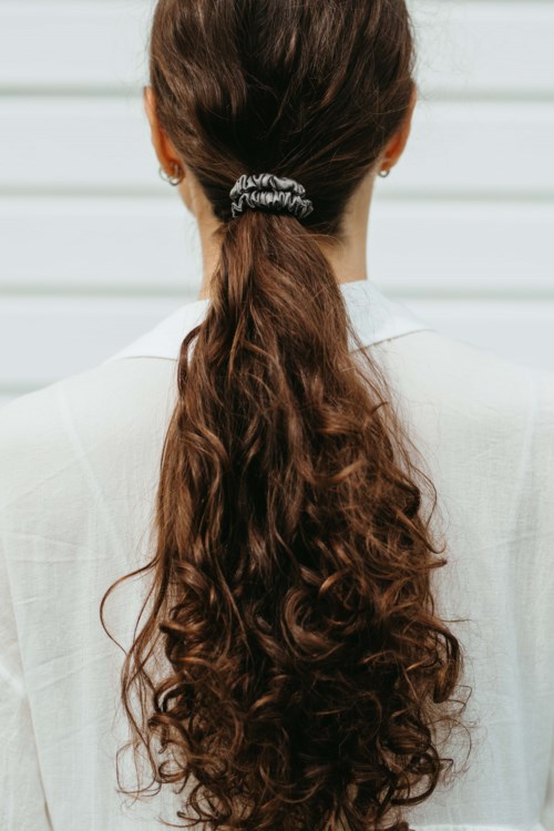 A woman with long, curly brown hair tied back with a hair tie, seen from behind. She wears a light-colored top.