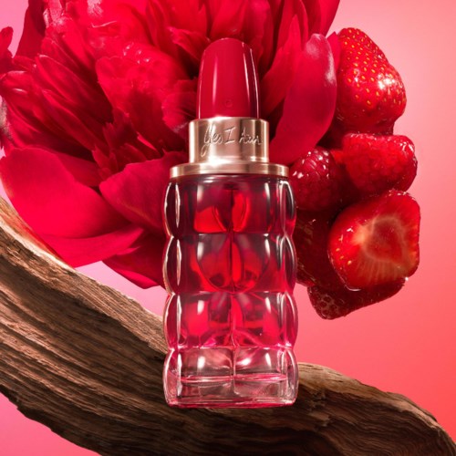 A perfume bottle with a red lipstick cap, surrounded by strawberries and pink flowers, on a wooden surface.