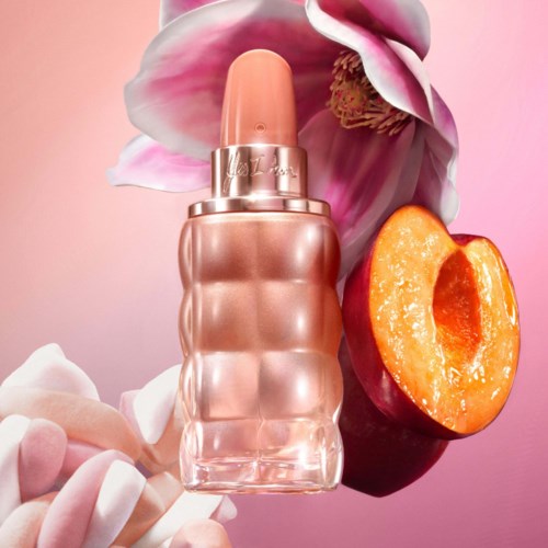 A perfume bottle surrounded by a flower, a peach half, and pink and white candy-like treats on a pink background.