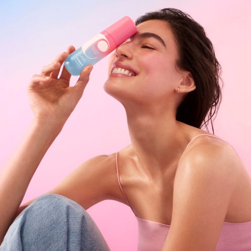 En smilende kvinne holder en rosa og blå sprayboks nær øyet, med en rosa bakgrunn.