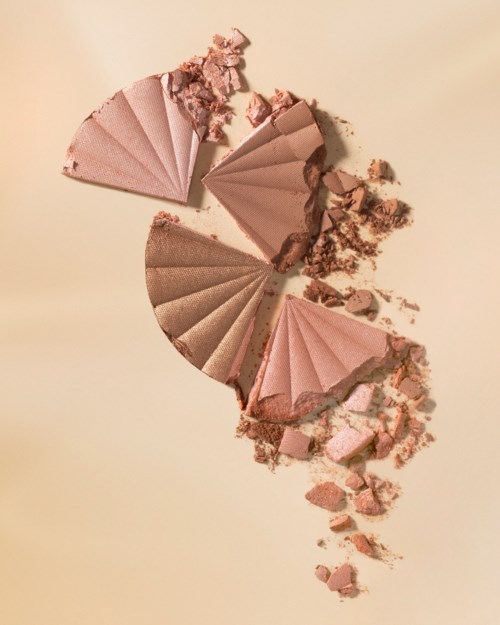 Gebroken make-up poeder in roze en beige tinten in waaiervorm met verspreide stukjes eromheen.