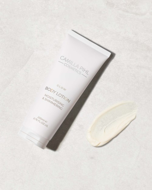 Een witte tube met 'Camilla Pihl Cosmetics Glow Body Lotion' erop en lotion naast het opgerold.