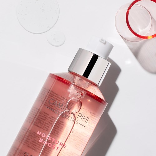 Een roze doorzichtige fles met een zilveren pomp, liggend op een witte ondergrond met een glas en waterdruppels ernaast.