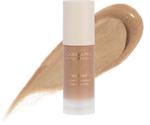 Eine Foundationflasche in Beige mit der Aufschrift 'Camilla Pihl Cosmetics Skin Tint Light Covering Foundation' und einem Probestrich dahinter.