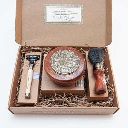 A shaving kit with a razor, shaving soap, and brush in a cardboard box with shredded paper.