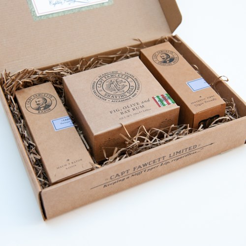 A cardboard box contains shaving soap bars with labels, placed on shredded packing material for protection.