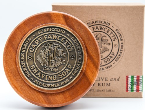 A round metal soap label inside a wooden dish, with a matching box behind. The label has detailed writing and a logo.