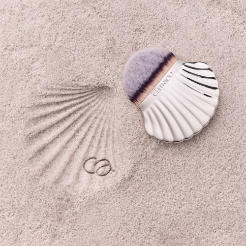 A makeup brush with a shell-shaped handle and the word 'Clarena' draws a wavy line in the sand, creating circles.