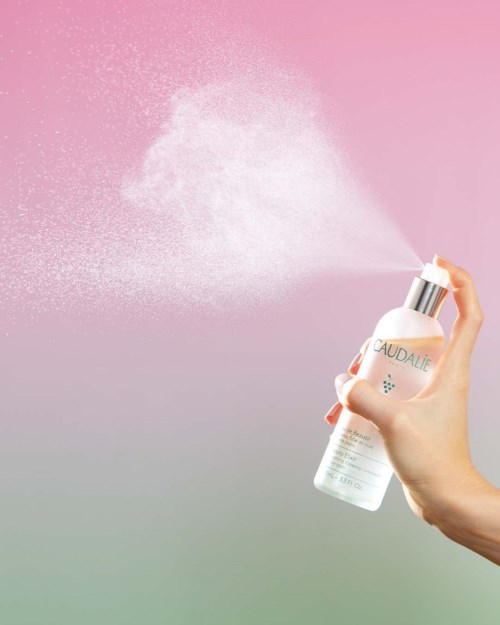 A hand sprays a white mist from a pump bottle against a pink and grey gradient background.
