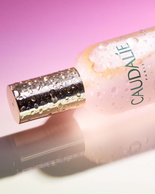 A bottle with a spray nozzle, pinkish liquid, and water droplets, labeled 'CAUDALIE PARIS' on a pink background.