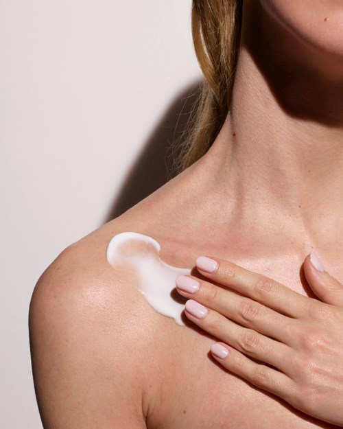 A woman applies lotion or cream on her shoulder, touching her collarbone with her hand. She has fair skin and light hair.
