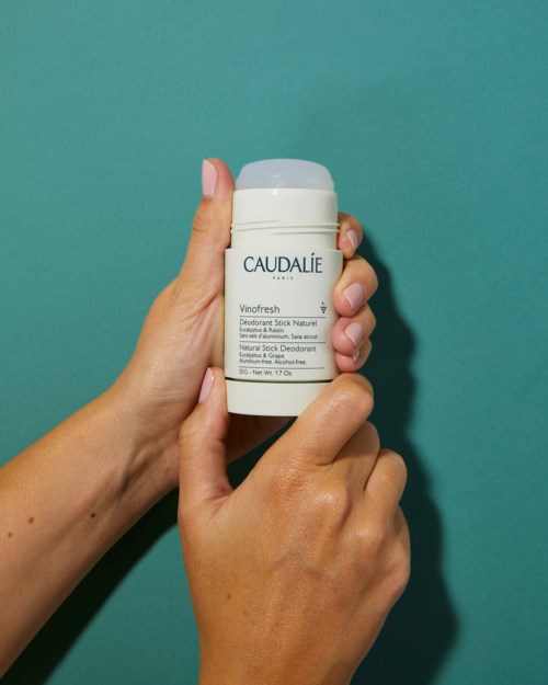 En hand håller en vit behållare märkt 'Caudalie Vinofresh deodorant stick' mot en turkost bakgrund.