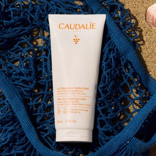 It is a white tube of after-sun lotion surrounded by a blue net, placed on sand.