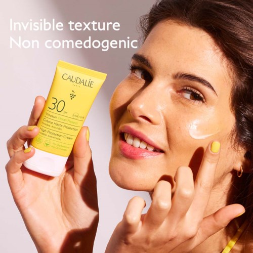 Nainen hymyilee ja pitää kädessään keltaista auringonsuojavoidetta merkinnällä 'Caudalie' ja SPF 30.