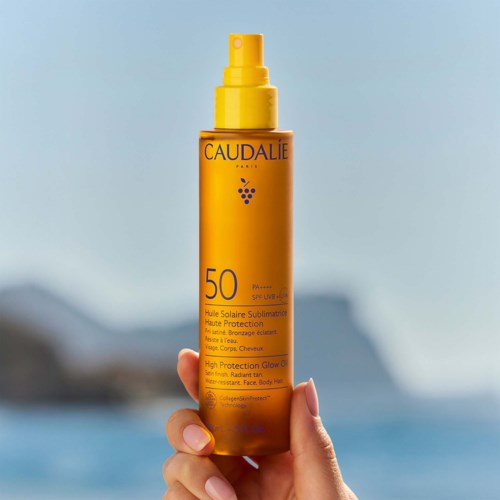 Käsissä on keltainen Caudalie aurinkosuoja-ruisku, SPF 50, ulkona veden lähellä.