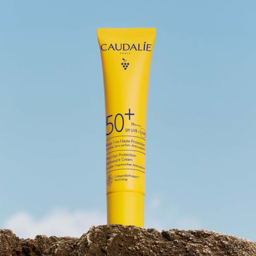 A yellow sunscreen tube with SPF 50+ stands on a rock, with a blue sky and ocean in the background.