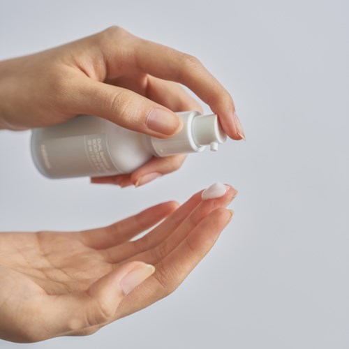 Eine Person gibt Lotion aus einer weißen Flasche auf ihre Hand, vor einfachem Hintergrund.