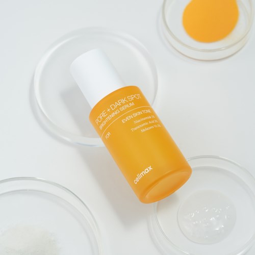 Orange flaska märkt Pore + Dark Spot, anti-fläck-serum, på vit yta med små skålar.