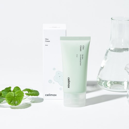 Eine grüne Tube Cica Cream mit Verpackung, ein Glas Wasser und grüne Blätter auf einer weißen Oberfläche.