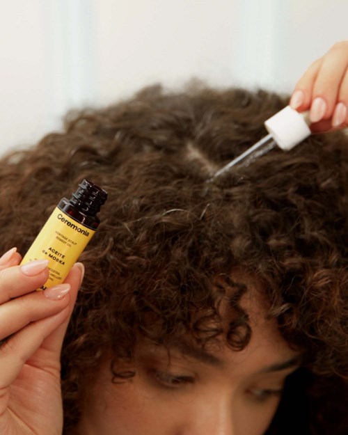 A person applies essential oil to curly hair using a dropper, with a bottle labeled 'Ceremonia' in hand.