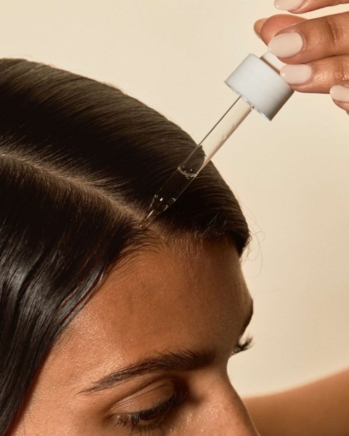 A woman uses a dropper to put oil into her hair part. She has long, dark, straight hair.