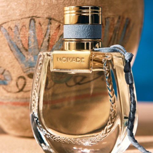 Een parfumfles met de naam NOMADE, met een gouden en blauwe dop, doorzichtig glas en een decoratief touw. Daarachter een bruine pot.