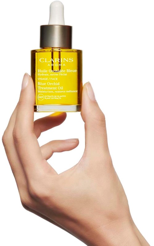Eine Hand hält eine gelbe Glasflasche mit Tropfer, beschriftet Clarins Blue Orchid Treatment Oil für das Gesicht.