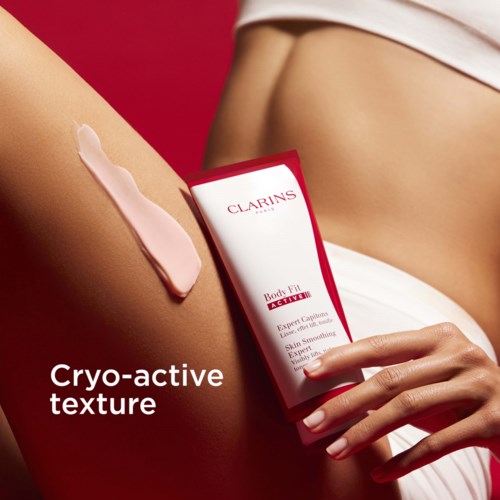 A woman applies pink cream to her thigh, holding a red and white box of Clarins skincare product.