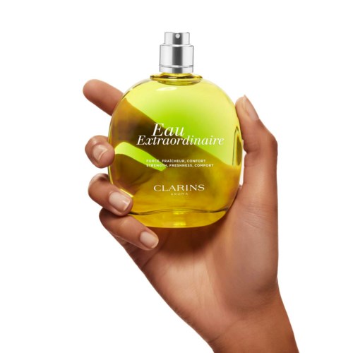 A hand holds a yellow perfume bottle with a spray top, labeled Eau Extraordinaire by Clarins.