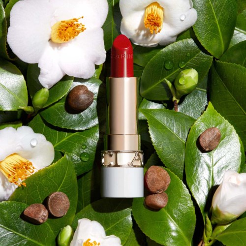 A red lipstick case surrounded by green leaves, white flowers, and small stones.