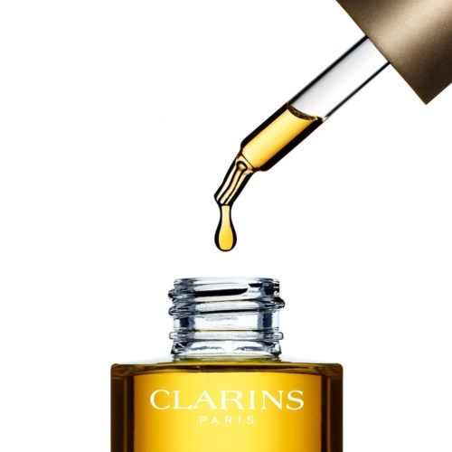 Eine Pipette tropft goldene Öl in eine kleine Flasche mit dem Label 'Clarins Paris' auf weißem Hintergrund.