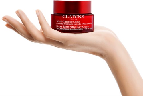 Käsi pitää punaista Clarins-voidepurkkia. Tausta on valkoinen ja musta.