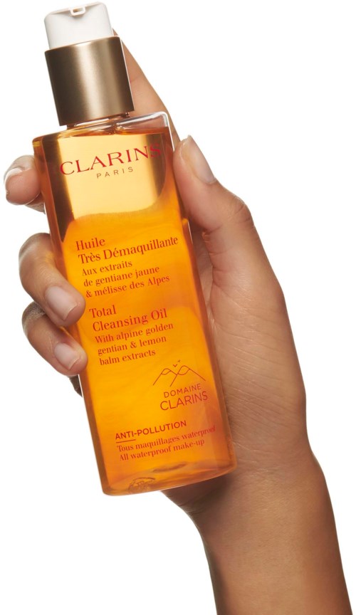 Kädessä on kirkas oranssi pullo, jossa on valkoinen pumpu ja etiketti Clarins Total Cleansing Oil Alpine Gold salts.