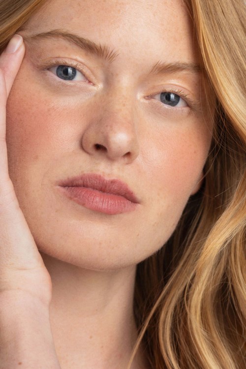 Close-up van een vrouw met rood haar, blauwe ogen en lichte huid, die haar gezicht voorzichtig aanraakt.