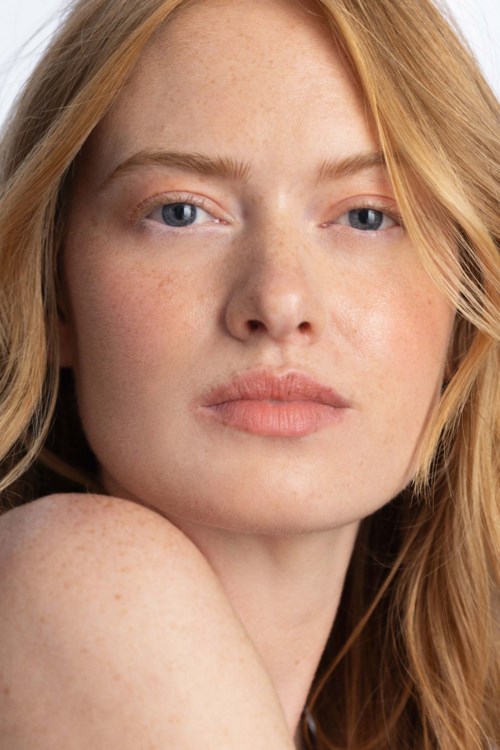 Een jonge vrouw met rood haar, lichte huid, blauwe ogen en natuurlijke make-up kijkt in de camera.