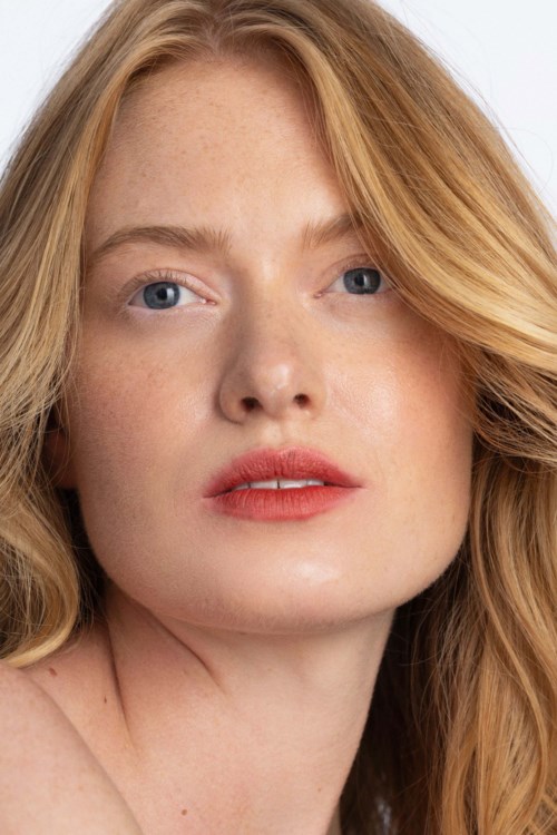 Een vrouw met rood haar en blauwe ogen heeft een neutrale uitdrukking, met zachte make-up en natuurlijke huid.