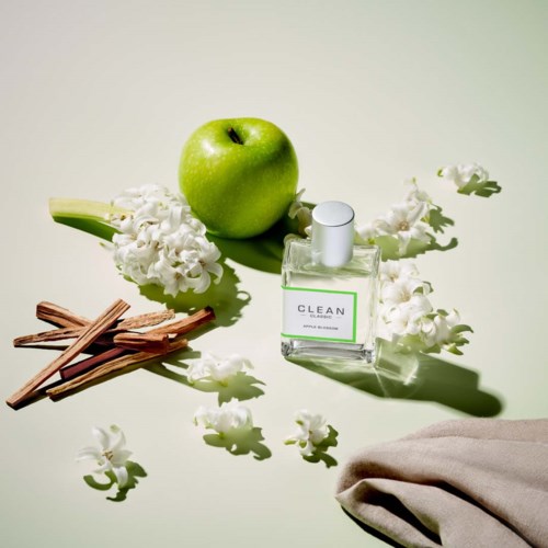 Ein grüner Apfel, weiße Blumen, Zimtstangen, Parfümflasche und graues Tuch auf hellem Hintergrund.