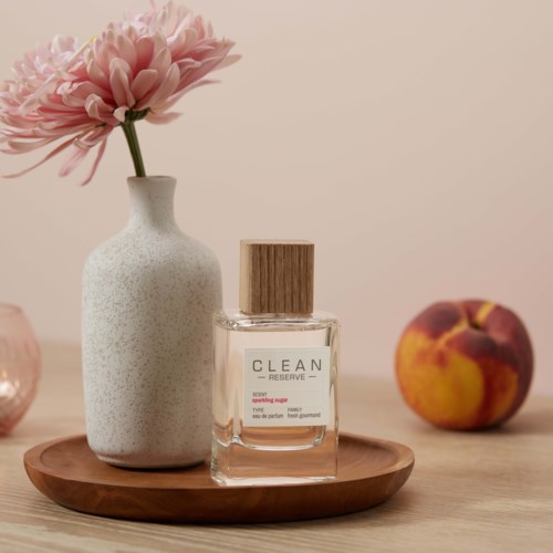 Eine weiße Vase mit einer pinken Blume, eine Glasparfümflasche mit Holzdeckel auf einem runden Holzteller mit einem Pfirsich.